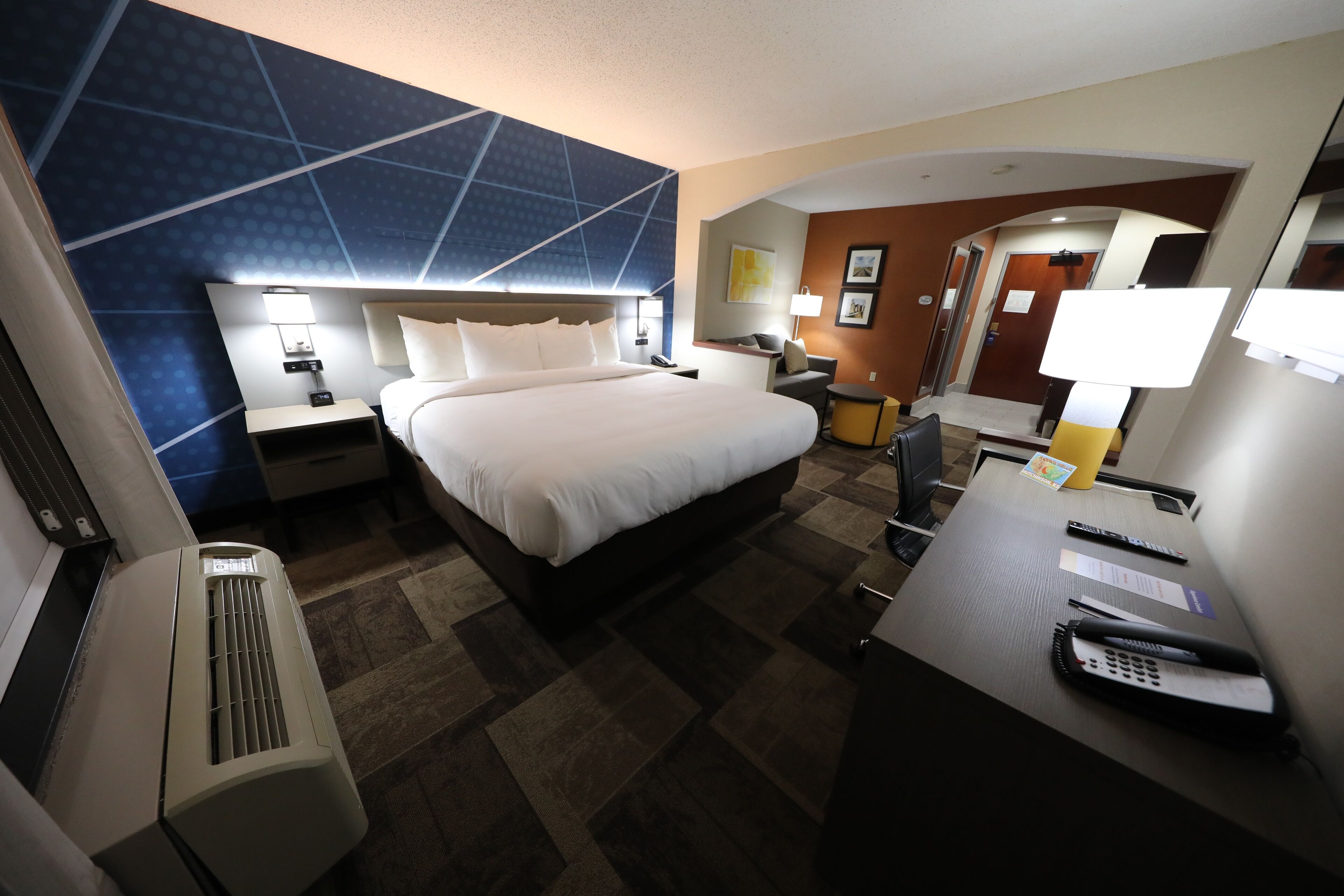 Habitación de hotel con una cama grande, un escritorio con teléfono, una silla y una lámpara de escritorio. Se observa un aire acondicionado de pared y un espejo.