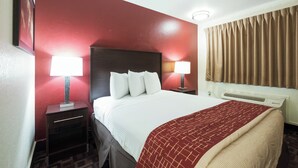 A hotel room with a large bed, two bedside tables with lamps, a red accent wall, a window with curtains, and a radiator. - Red Roof Inn Oklahoma Airport – I-40 W/Fairgrounds (Oklahoma City)
