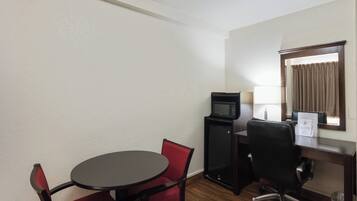 Ein Hotelzimmer mit einem runden Tisch, zwei roten Stühlen, einem schwarzen Schreibtisch mit Computer und Mikrowelle, einem weiteren Stuhl, einem Fenster mit Vorhängen und einem Mülleimer.