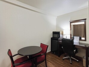 A hotel room with a round table, two red chairs, a black desk with a computer and a microwave, a chair, a window with curtains, and a trash bin. - Red Roof Inn Oklahoma Airport – I-40 W/Fairgrounds (Oklahoma City)