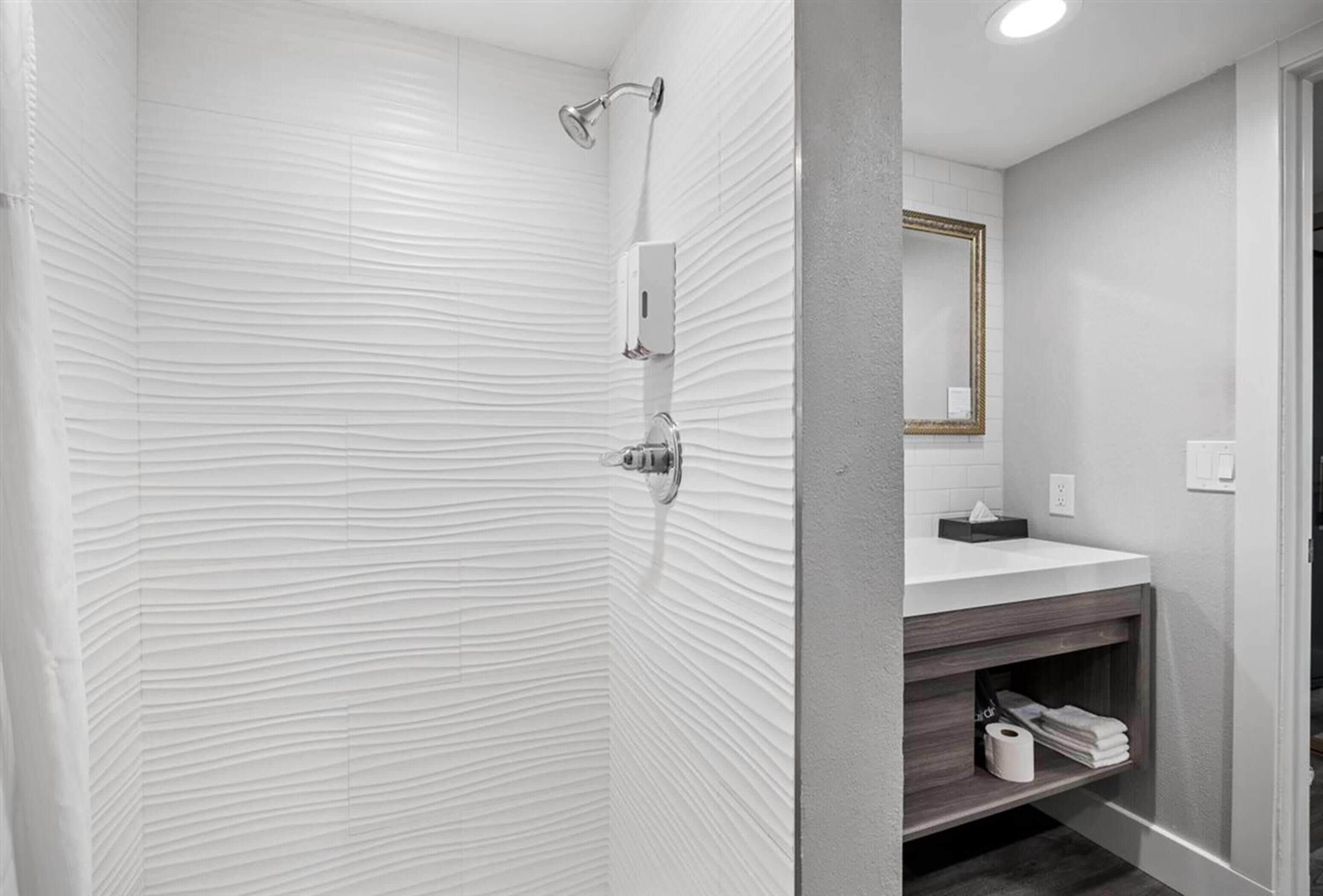 A modern bathroom with a textured white wall, a shower with a handheld showerhead, a vanity with a mirror, and a shelf with towels.