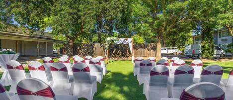Rows of white chairs with red sashes, arranged in a semi-circle outdoors with trees and a building in the background.