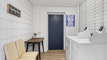 Una sala de lavandería con un cartel que dice "Laundry Room", una mesa con sillas, una puerta oscura y un panel de control en la pared.