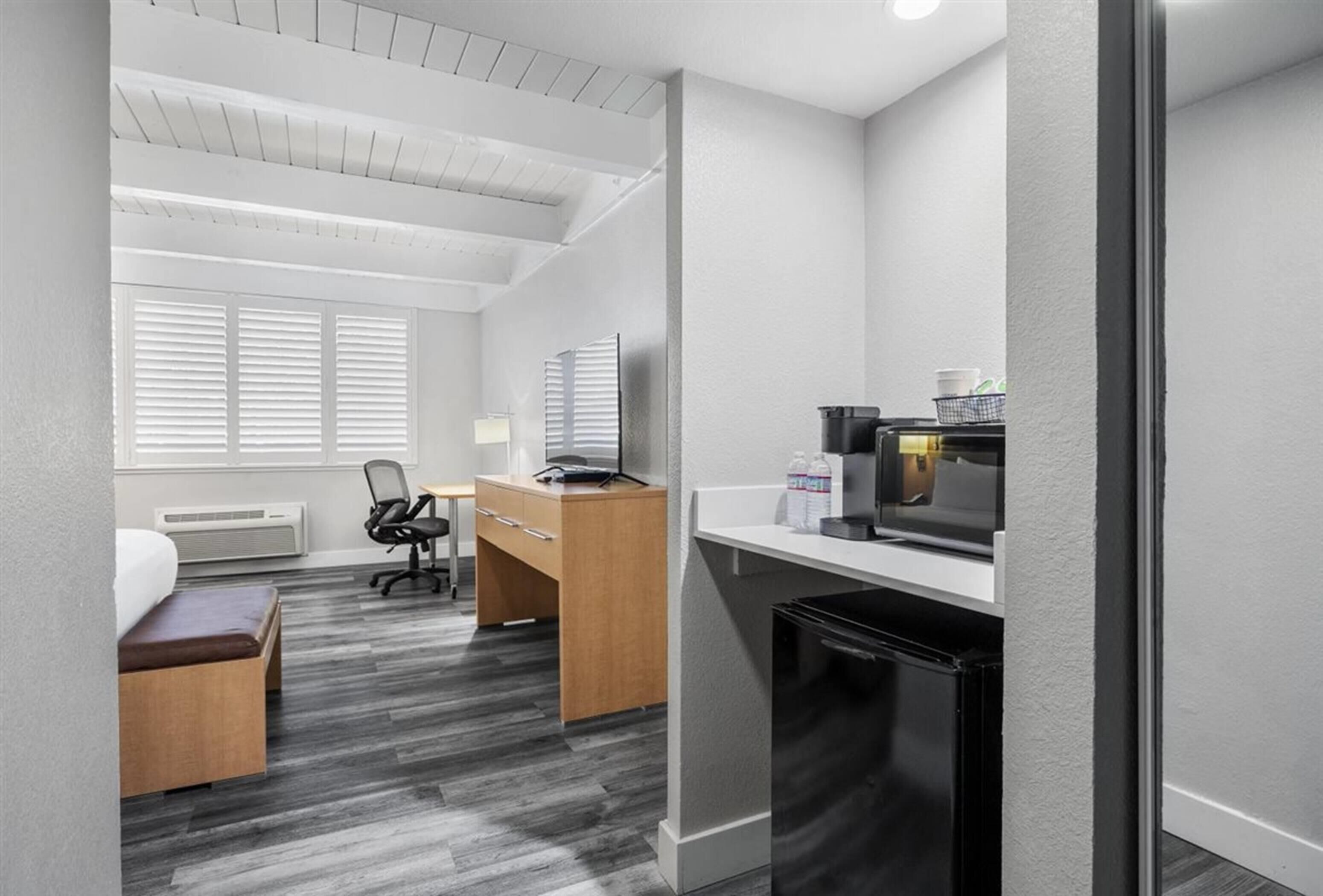 A compact hotel room with a kitchenette, a desk, a chair, a bed, and a window with blinds.