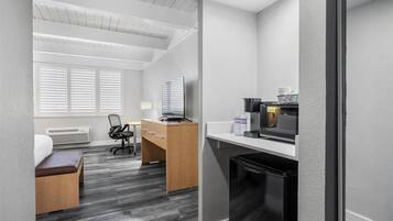 A compact hotel room with a kitchenette, a desk, a chair, a bed, and a window with blinds.