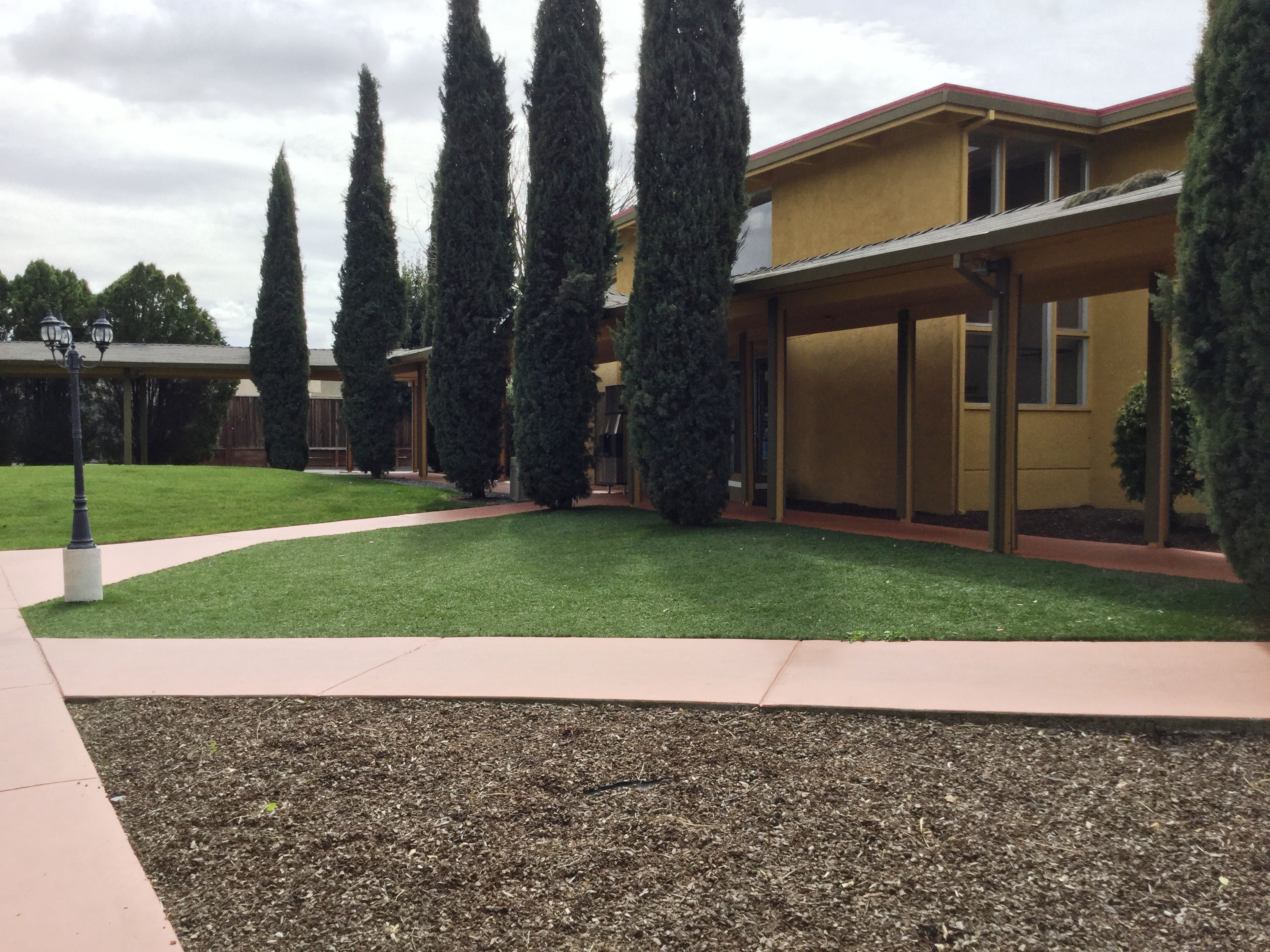 Un bâtiment à la façade jaune, doté d’un patio couvert et d’une pelouse bien entretenue, agrémentée d’arbres hauts et élancés.