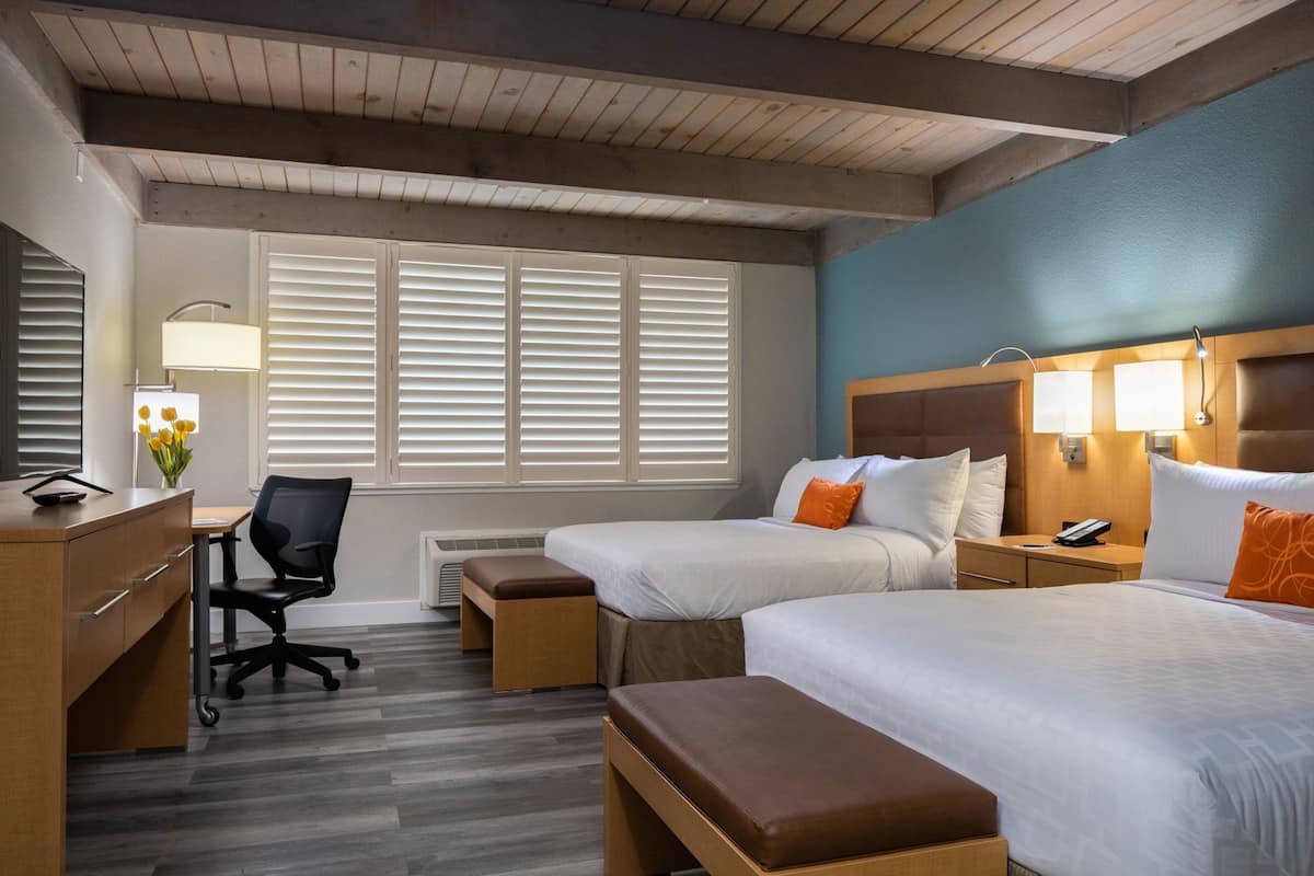 A hotel room with two beds, a desk, a chair, a lamp, and a window with blinds.