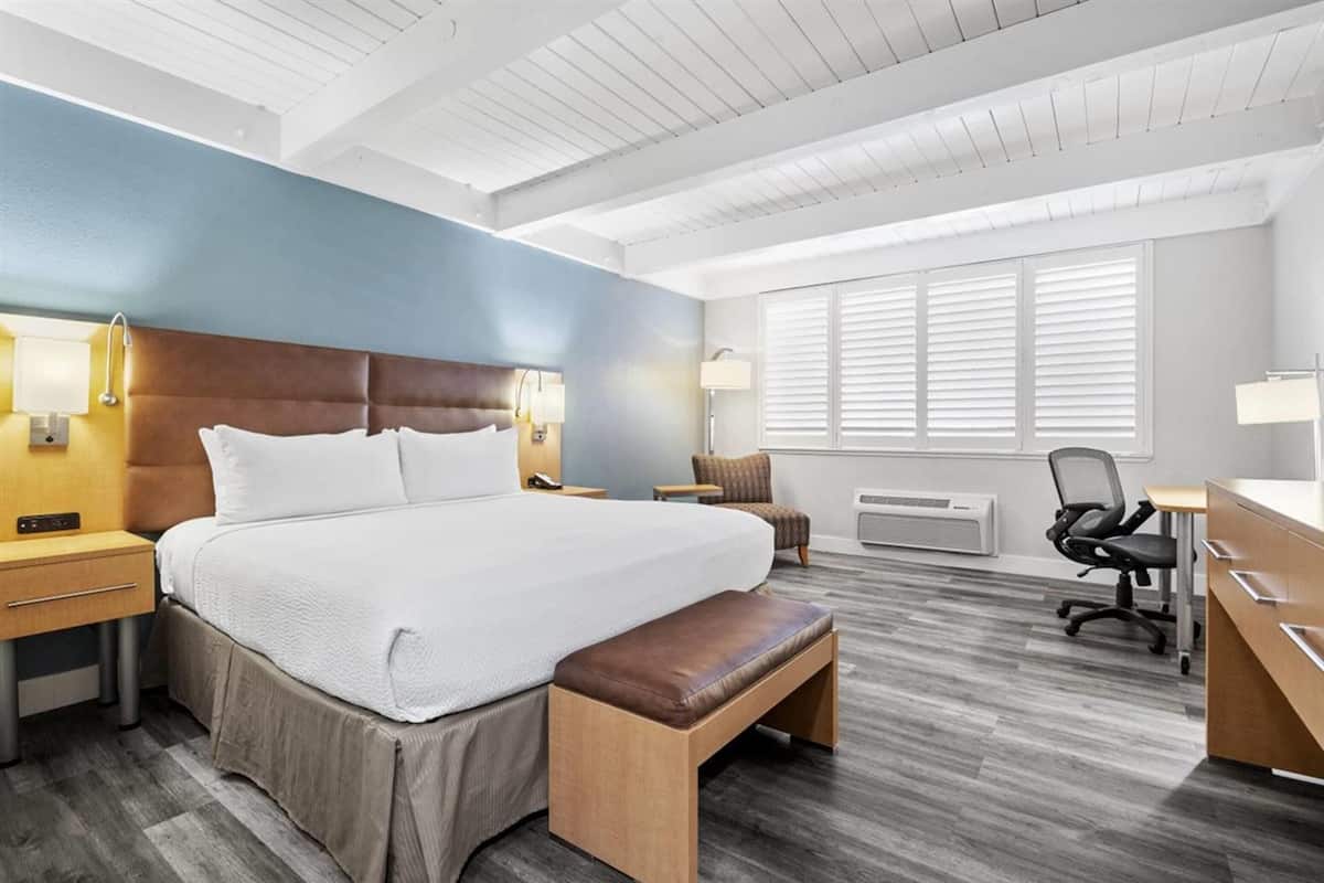 A hotel room with a large bed, a desk, a chair, and a bench. There are two bedside tables with lamps, a window with blinds, and a ceiling with wooden beams.