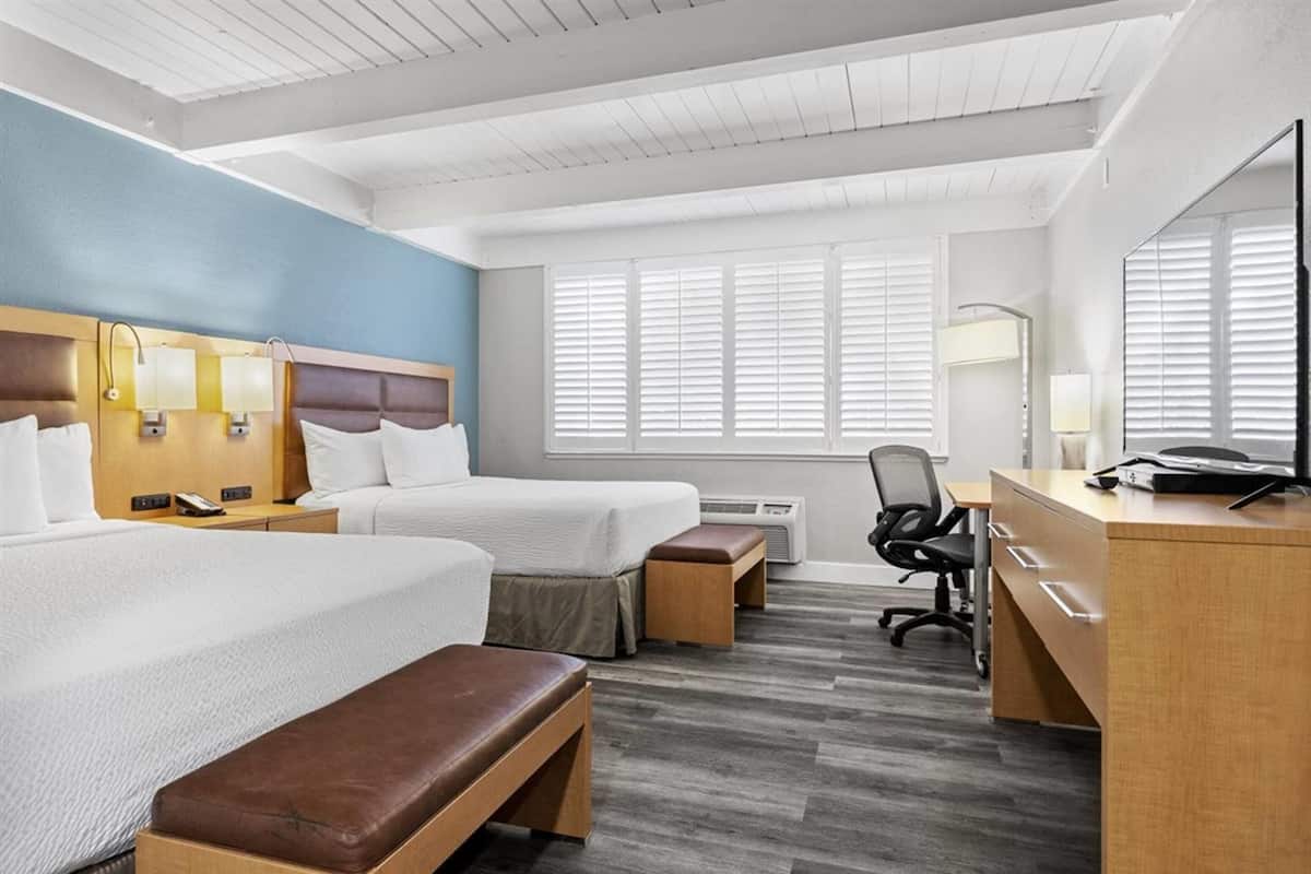 A hotel room with two beds, a desk, a chair, and a large window with blinds.