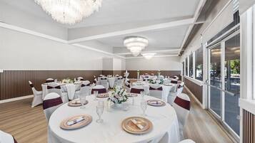 Un salón de banquetes con mesas redondas dispuestas para un evento formal, con candelabros y ventanas amplias.
