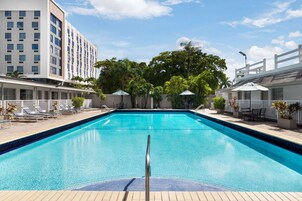 Una piscina con sillas de descanso y sombrillas, rodeada por un edificio de varios pisos y balcones.