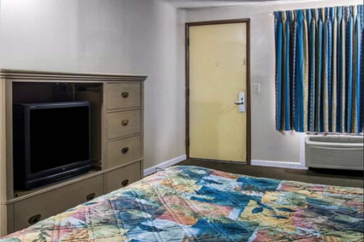 Hotellihuone, jossa on sänky, lipasto, televisio, ovi ja verhot.