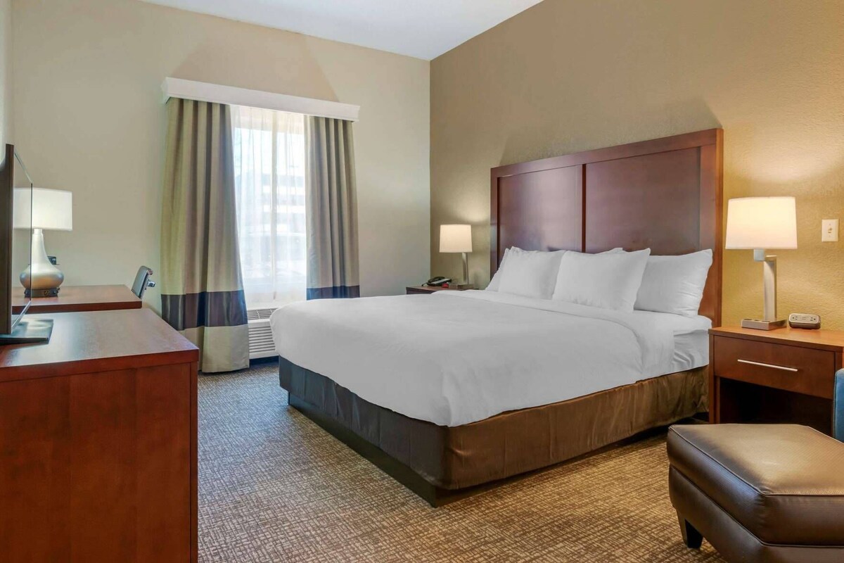 Ein Hotelzimmer mit einem großen Bett, einem Schreibtisch, einem Stuhl, einem Nachttisch mit Lampe und einem Fenster mit Vorhängen.