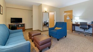 A hotel room with a blue sofa, a brown ottoman, a blue armchair, a desk with a chair, a television, and a mirror.
