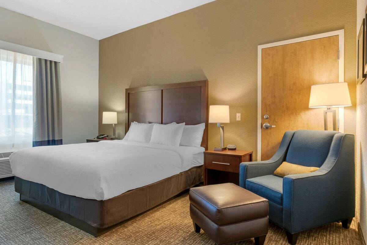 Une chambre d’hôtel avec un grand lit, une table de chevet avec une lampe, une chaise et un pouf.