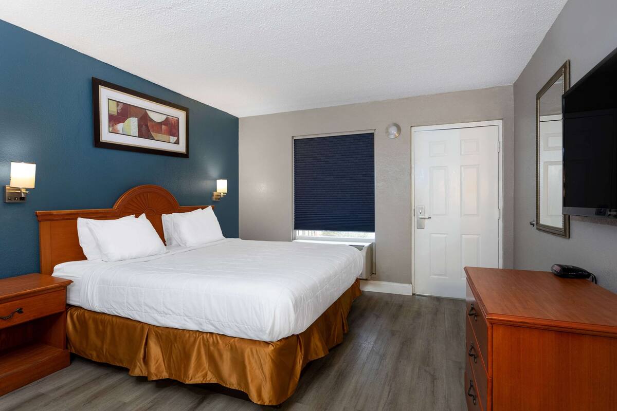 Une chambre d’hôtel avec un grand lit, une table de chevet en bois, une commode, un tableau encadré au mur et une fenêtre avec des stores.