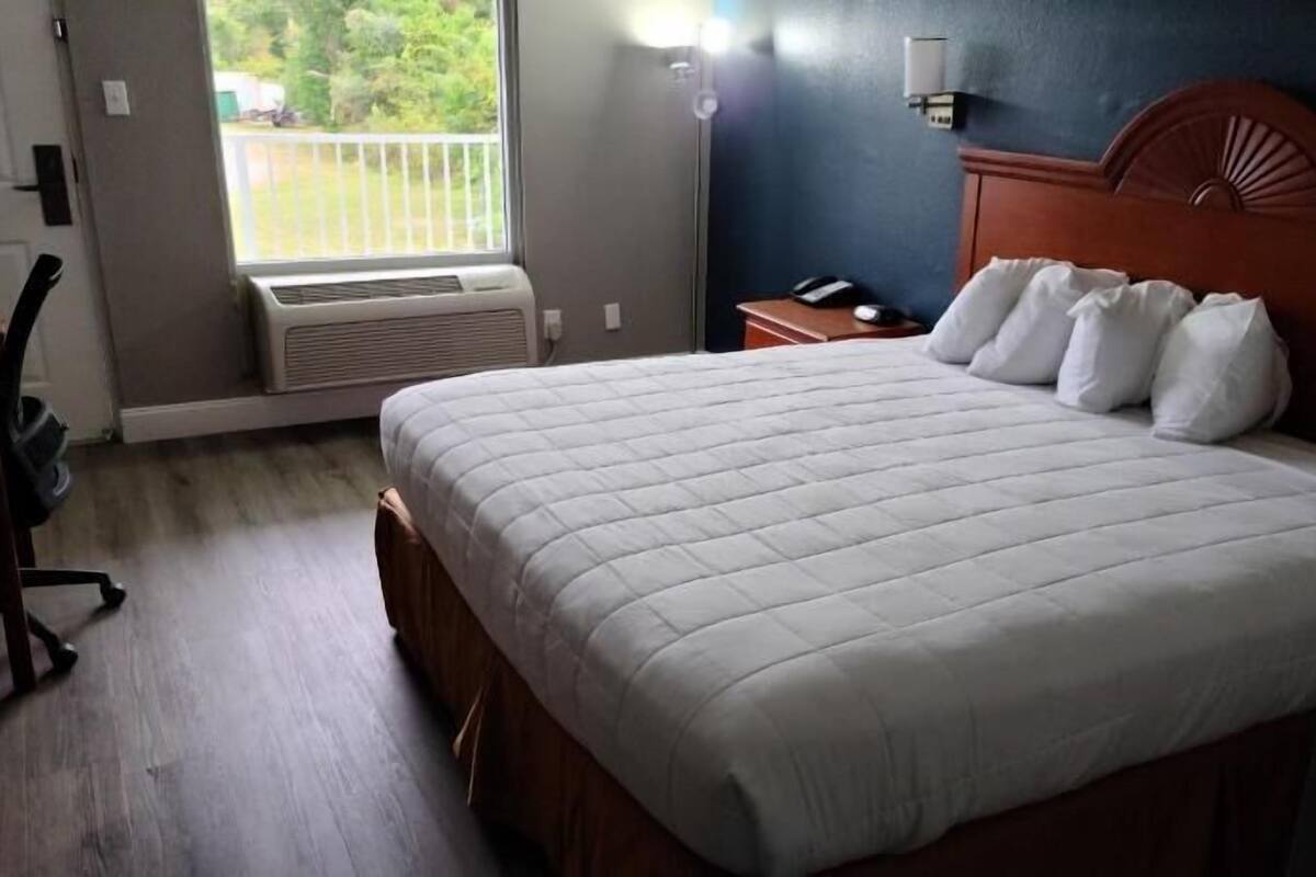 Une chambre d’hôtel équipée d’un lit, d’un bureau, d’une chaise, d’une table de chevet, d’une lampe et d’une fenêtre donnant sur un paysage verdoyant.