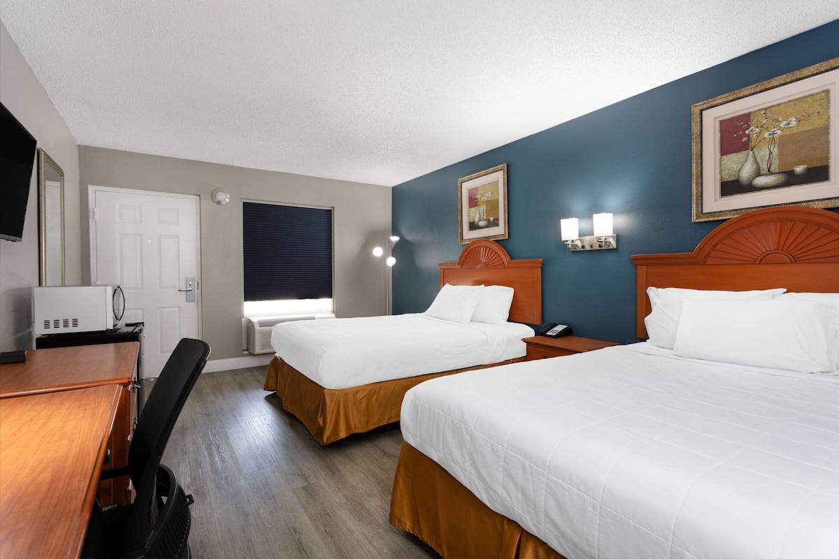 Une chambre d’hôtel avec deux lits, un bureau, une chaise, une télévision, une fenêtre avec des stores et des lampes fixées au mur.