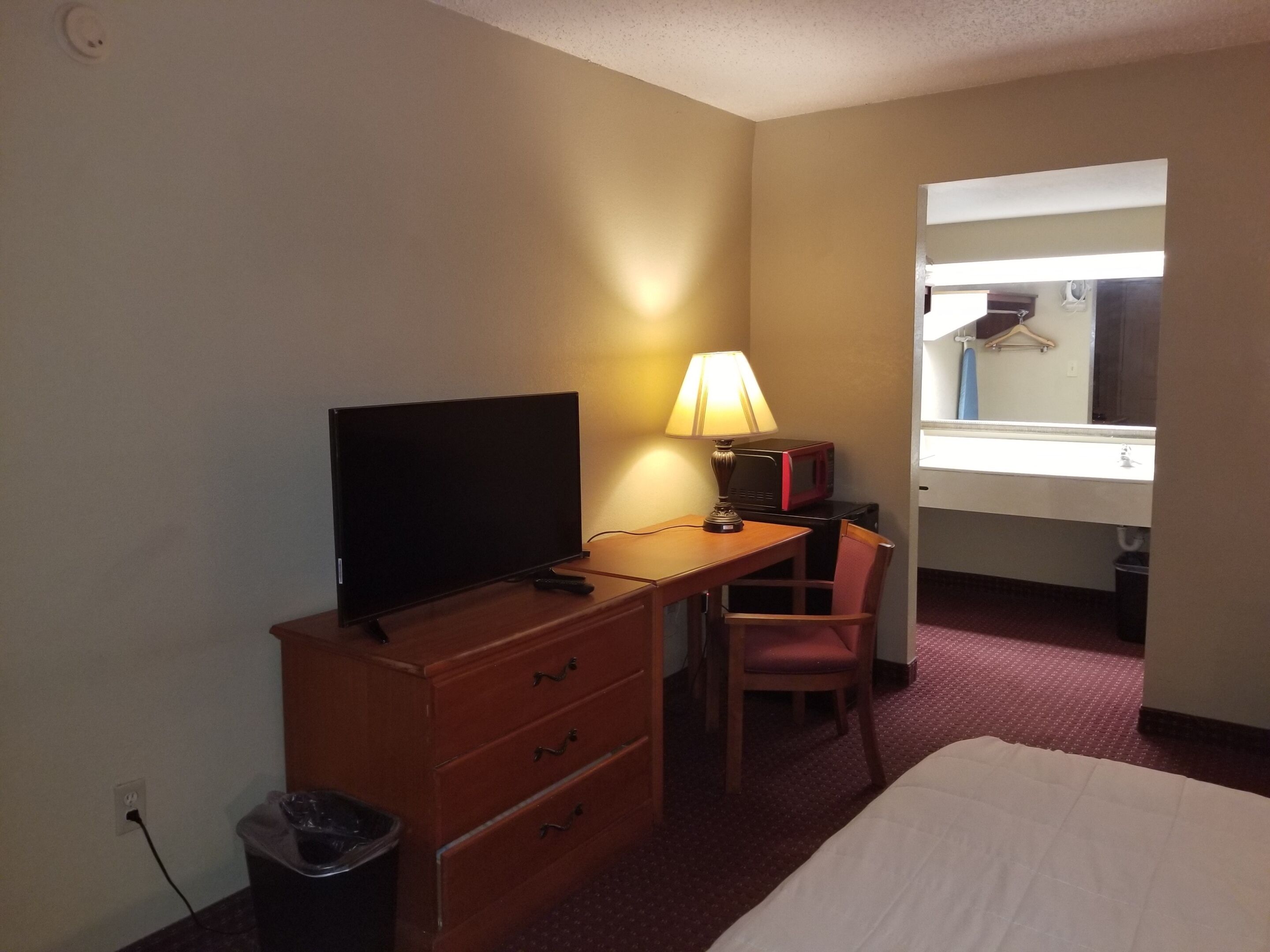 A hotel room with a bed, a desk with a TV, a lamp, a chair, a mirror, and a window.