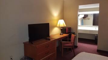 A hotel room with a bed, a desk with a TV, a lamp, a chair, a mirror, and a window.