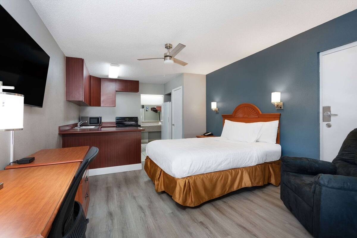 A hotel room with a bed, a desk, a kitchenette, a ceiling fan, and a TV.