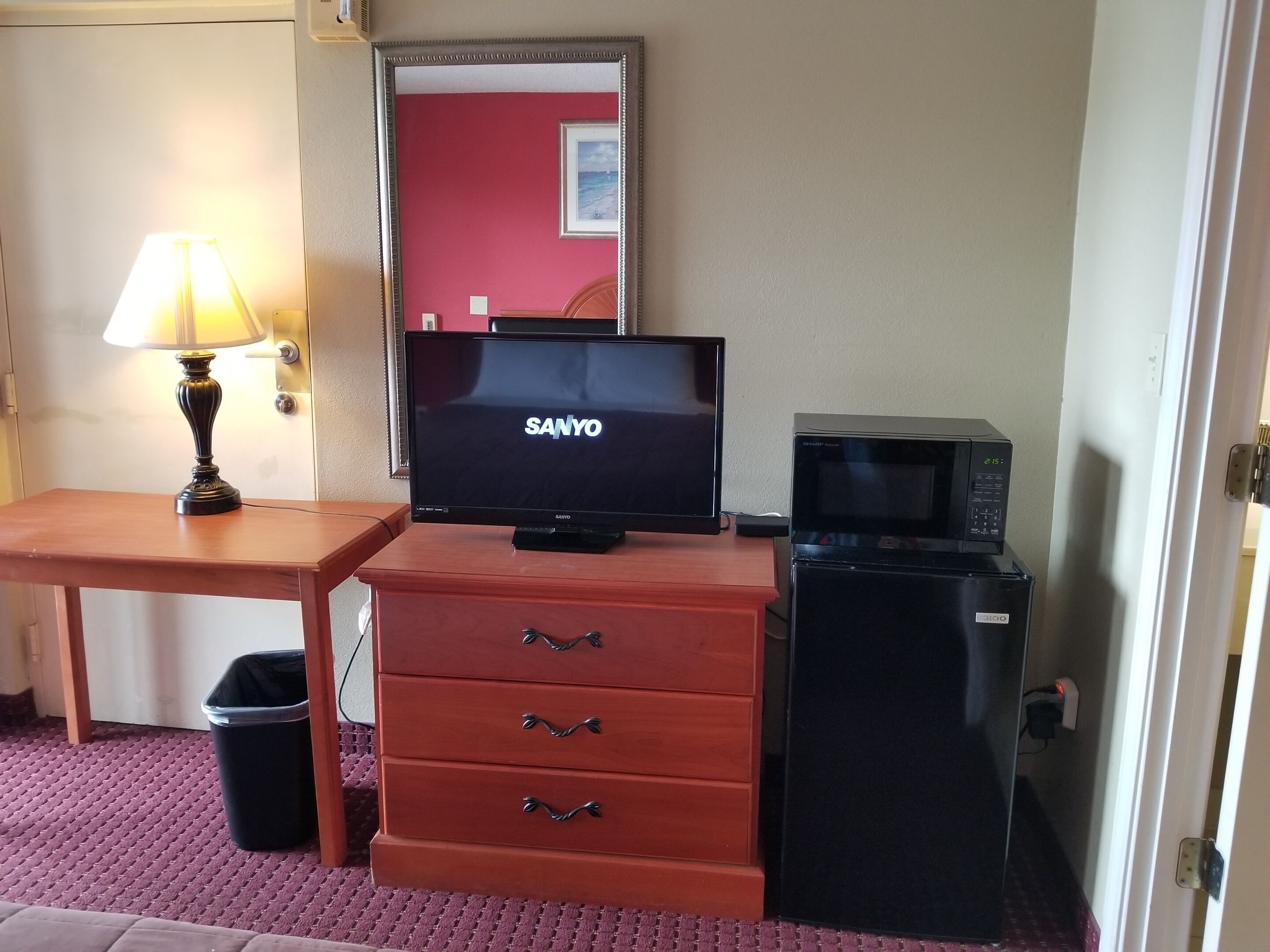 A hotel room with a television on a wooden dresser, a lamp on a table, a trash bin, and a microwave.