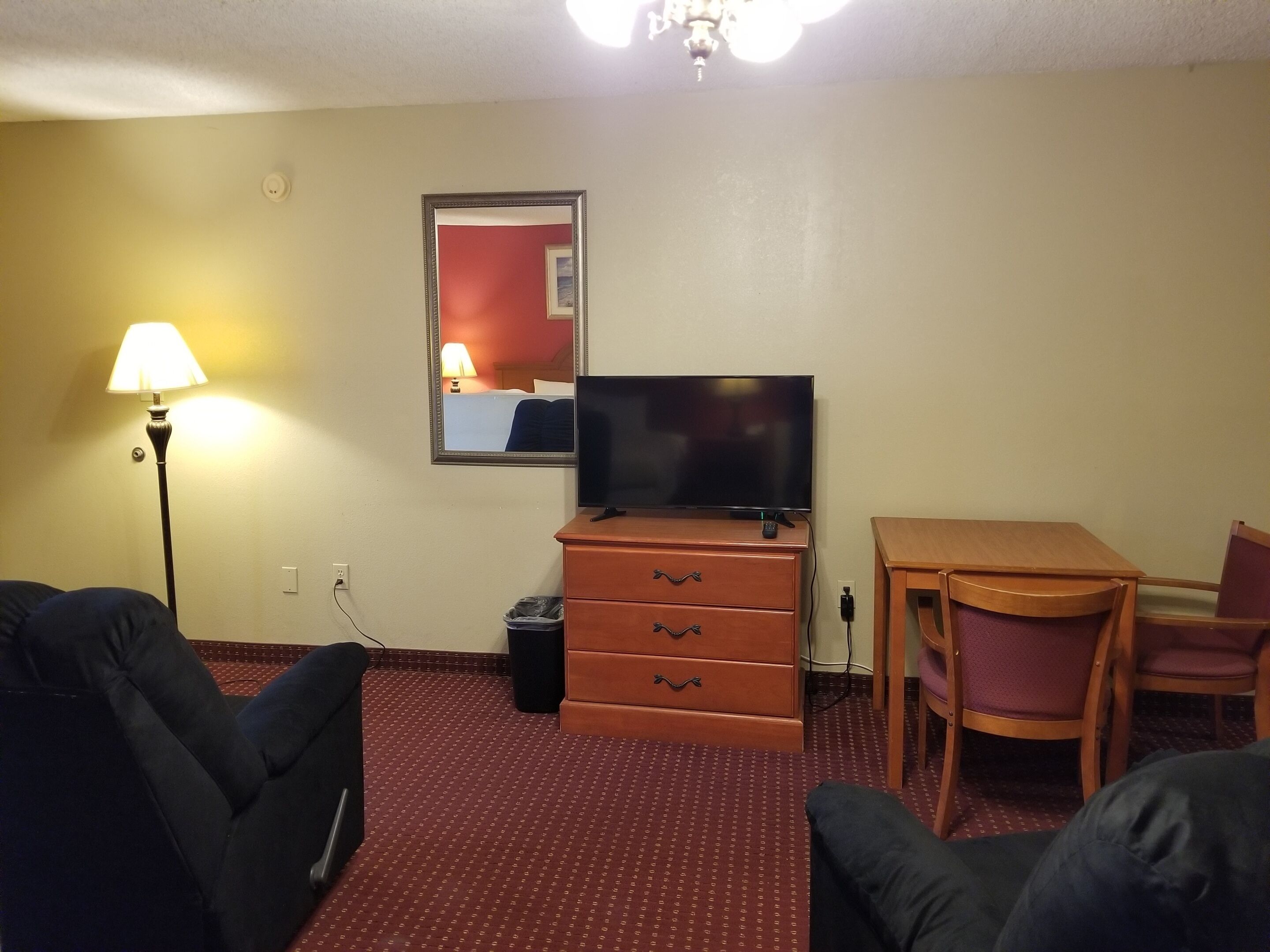 A hotel room with a TV on a dresser, a lamp, a dining table with chairs, and a carpeted floor.