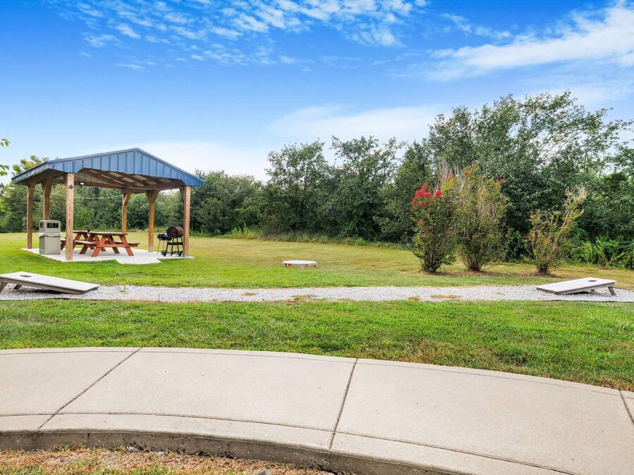 A gazebo with picnic tables and a barbecue grill in a grassy area with trees and a clear sky.