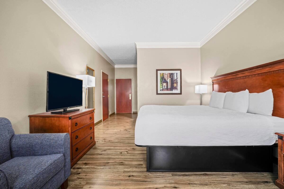 Habitación de hotel con cama, televisor sobre una cómoda de madera, una silla y un pasillo que conduce a otra habitación.