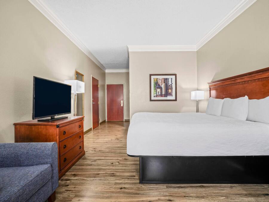 Une chambre d’hôtel avec un lit, un téléviseur posé sur une commode en bois, une chaise et un couloir menant à une autre pièce.