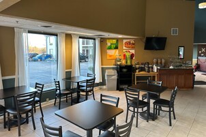 Interior de restaurante con mesas y sillas, una barra con dispensador de bebidas, y vista al área de estacionamiento a través del ventanal.