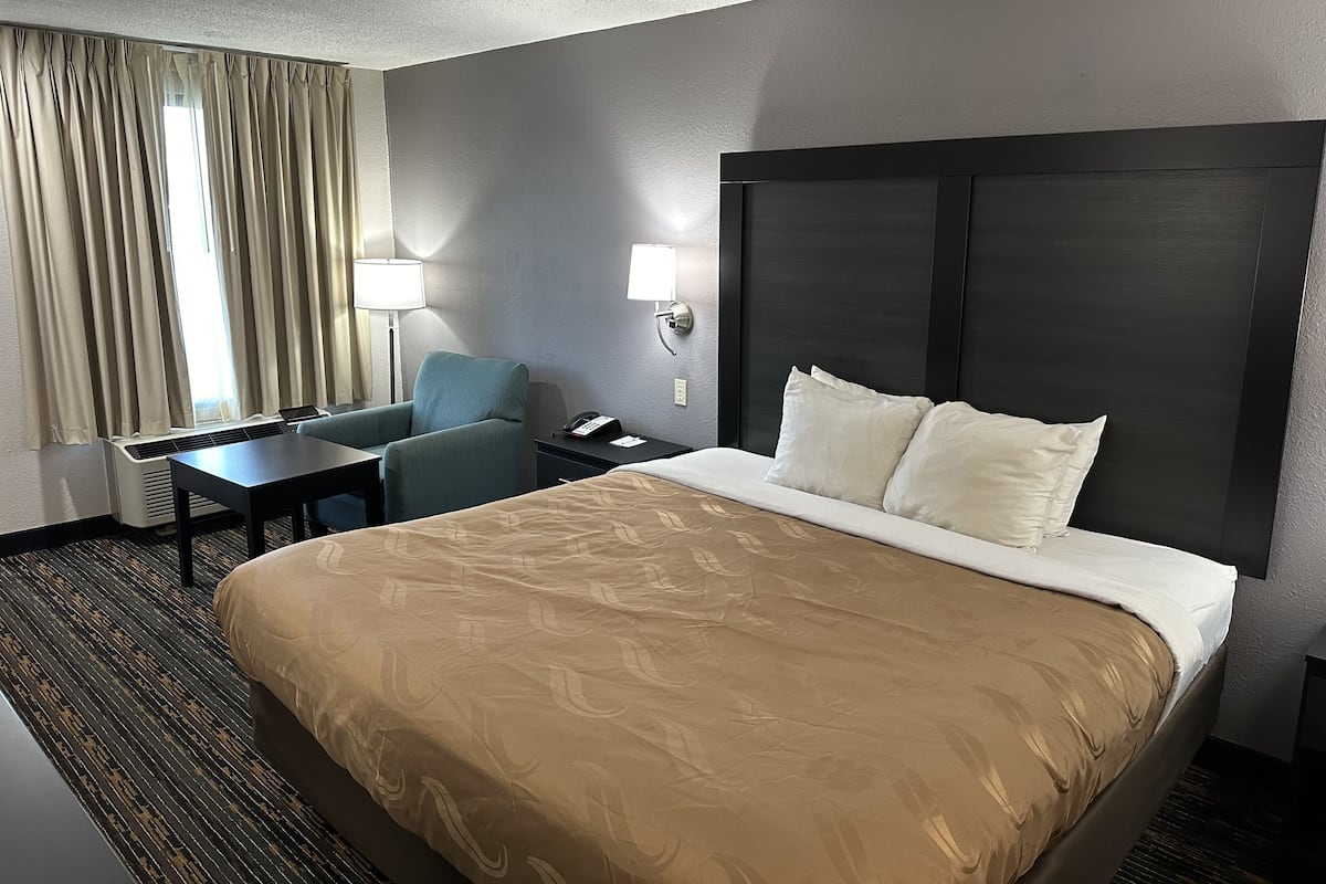 A hotel room with a large bed, a desk, a chair, a lamp, and a window with curtains.