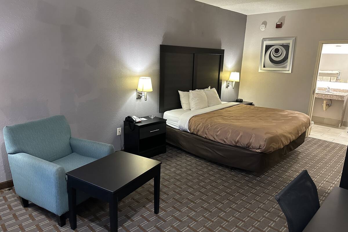 A hotel room with a bed, bedside tables, a chair, a small table, and a wall-mounted lamp.