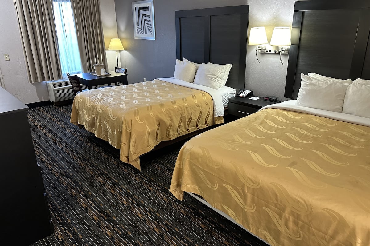 A hotel room with two beds, a desk, a chair, a lamp, and a wall-mounted phone.