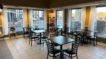 Een restaurantinterieur met meerdere tafels en stoelen, grote ramen met gordijnen en uitzicht op de straat.