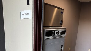 A vending machine labeled "ICE CUBE DISPENSER" in a hallway with wooden flooring and beige walls.