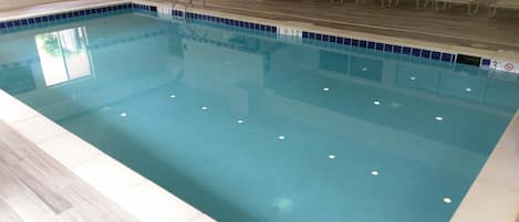 Una piscina interior con agua cristalina, una terraza de madera y sillones de descanso a lo largo de la pared.