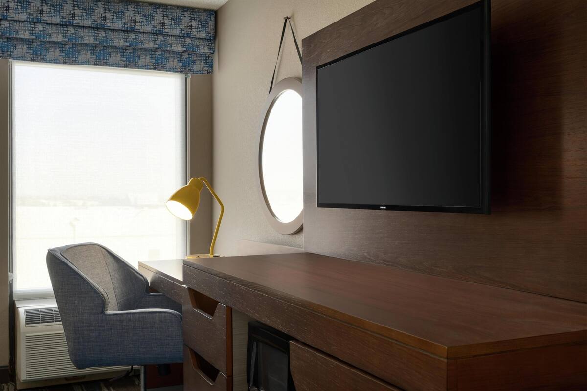 A hotel room with a wooden desk, a chair, a TV mounted on the wall, a window with a view, and a lamp.