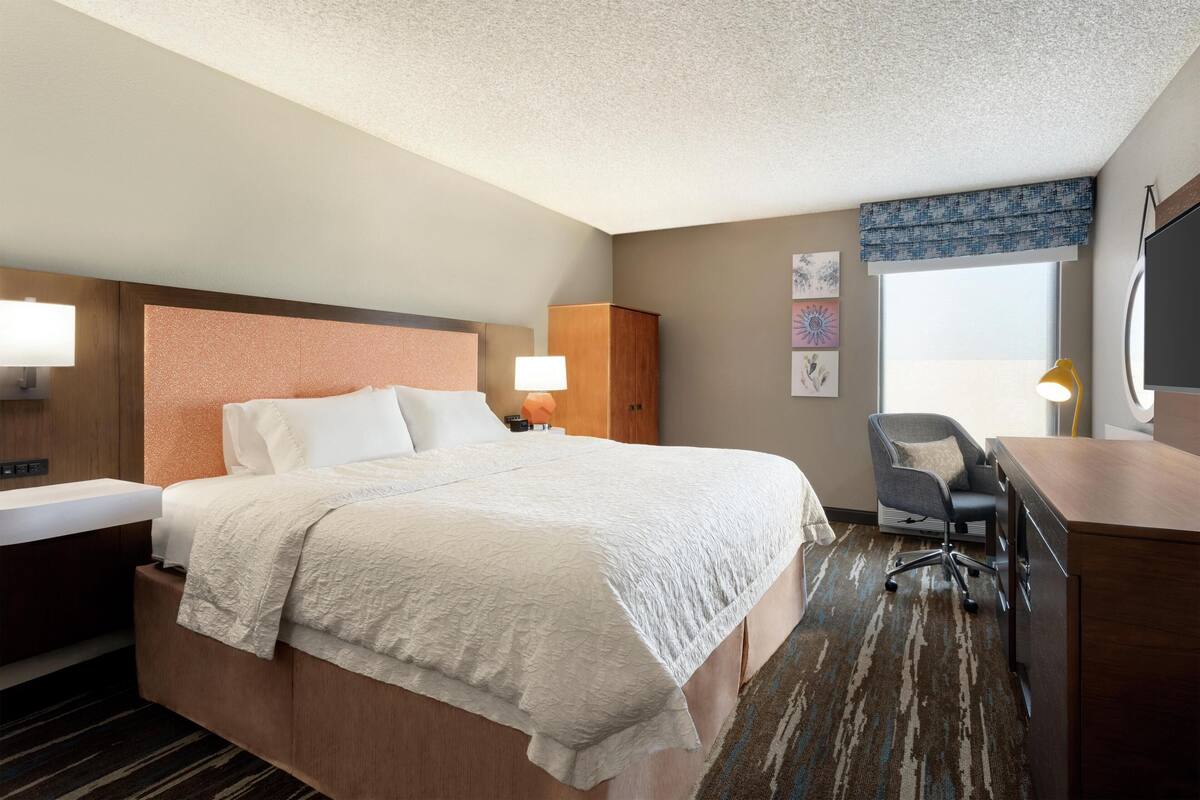 A hotel room with a large bed, a desk, a chair, a wardrobe, and a window with a view.