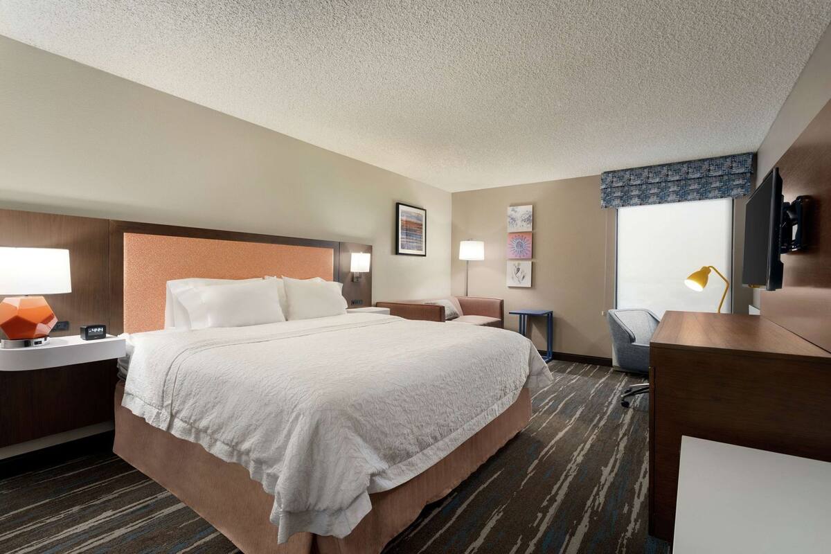 A hotel room with a large bed, bedside tables, a desk, a chair, a TV, and artwork on the walls.