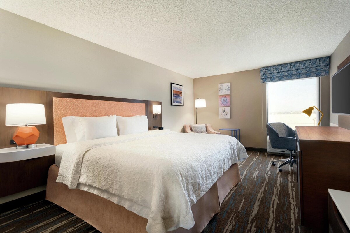 A hotel room with a large bed, a desk, a chair, a nightstand with a lamp, and a window with a view.