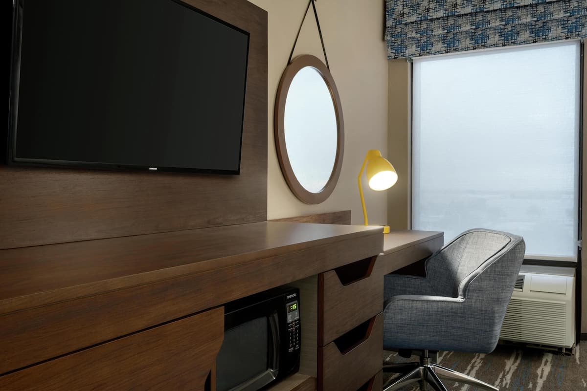 A hotel room with a wooden desk, a television, a round mirror, a desk lamp, a microwave, a chair, and a window with a view.