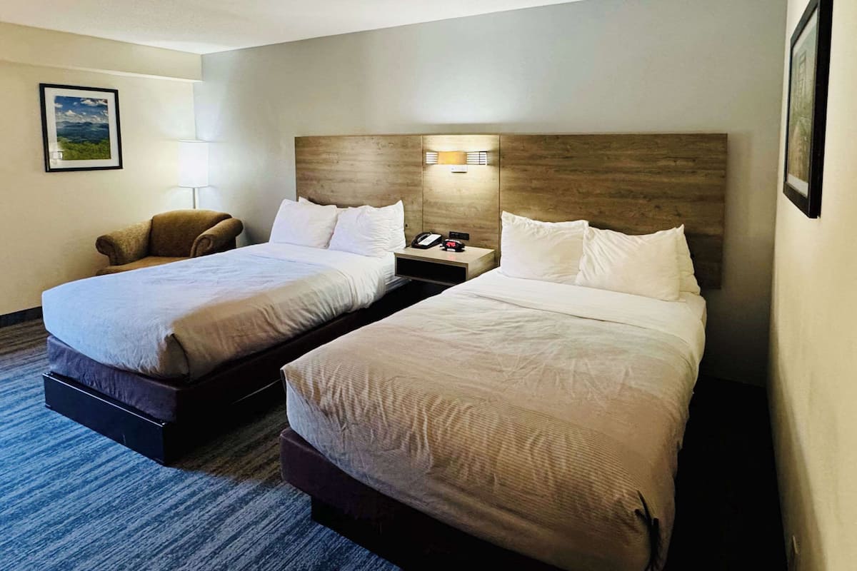Quarto de hotel com duas camas, mesa de cabeceira com luminária, uma cadeira e um quadro na parede.