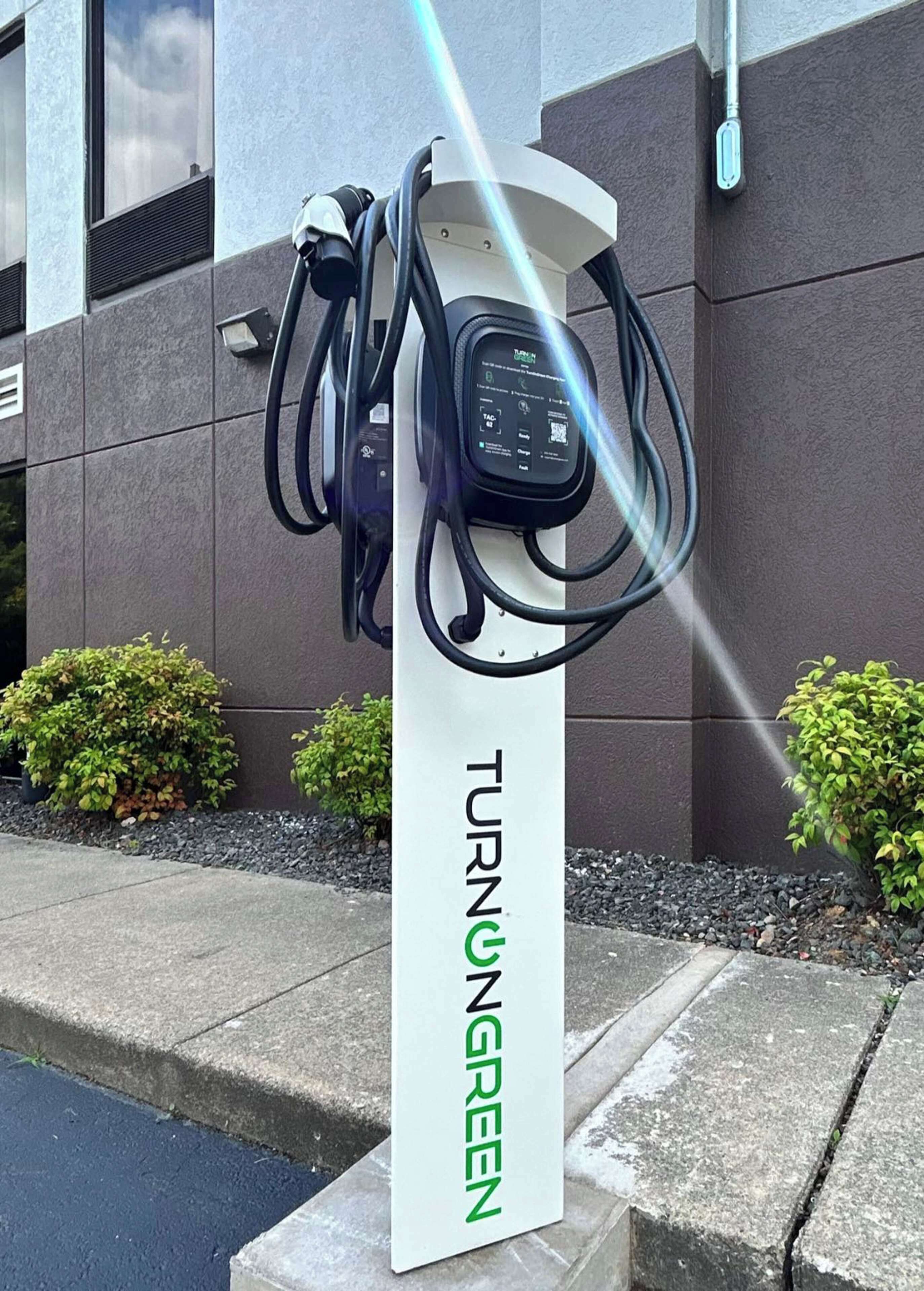 An electric vehicle charging station with a sign reading "TURNING GREEN" in front of a building with a grey facade and small bushes.