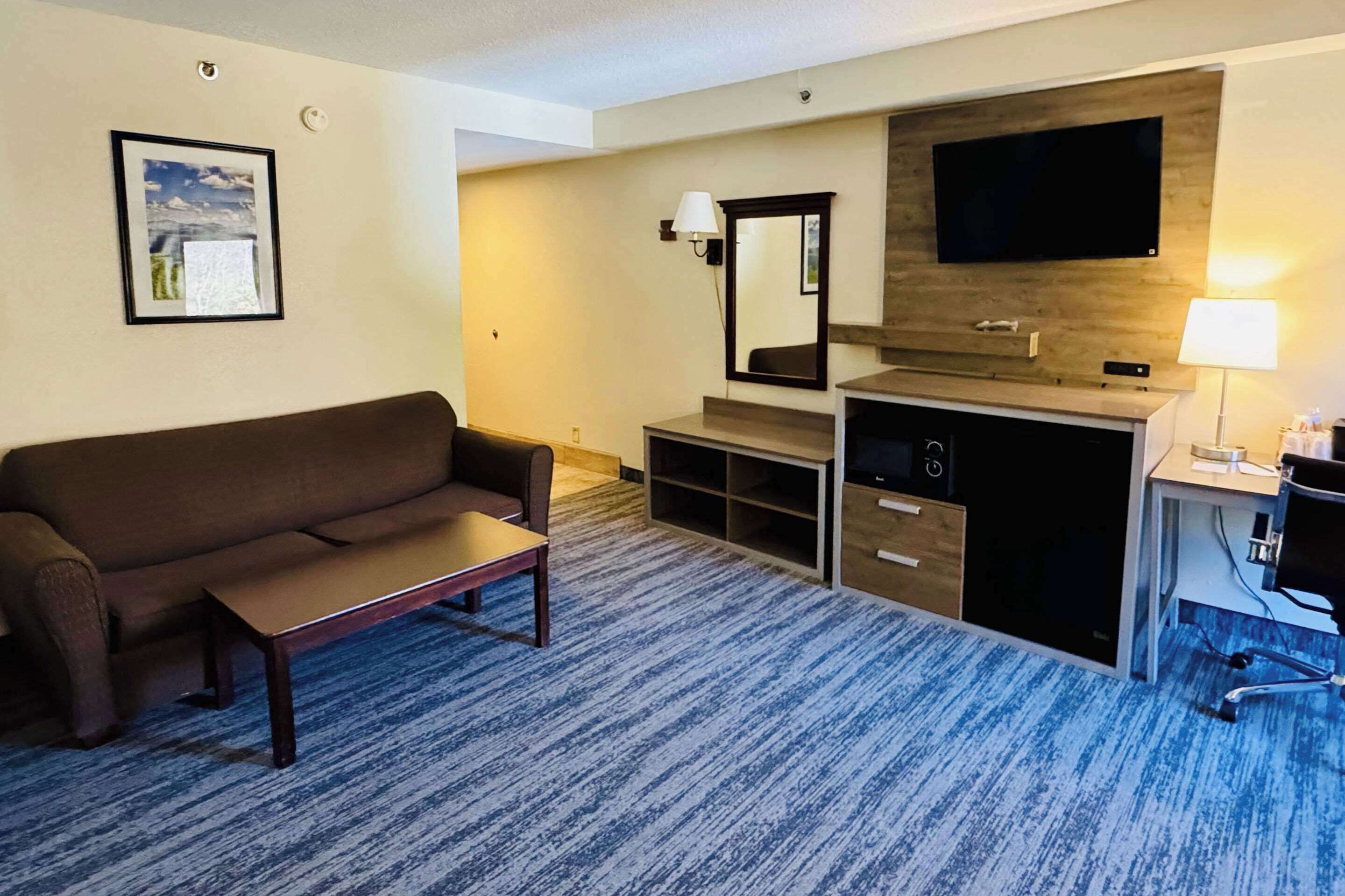 A hotel room with a sofa, a small table, a desk with a chair, a TV mounted on the wall, and a lamp.