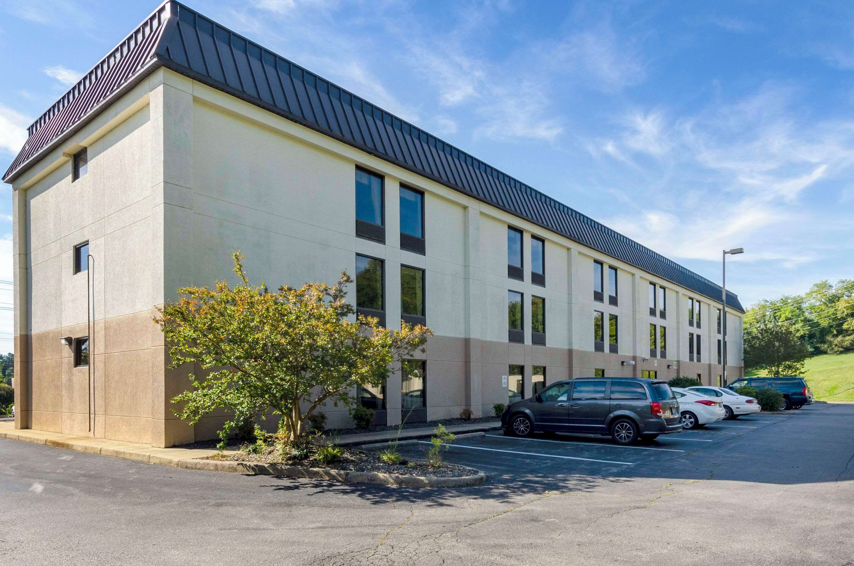 Un edificio moderno con techo plano y amplias ventanas. Se observa un estacionamiento con varios automóviles.
