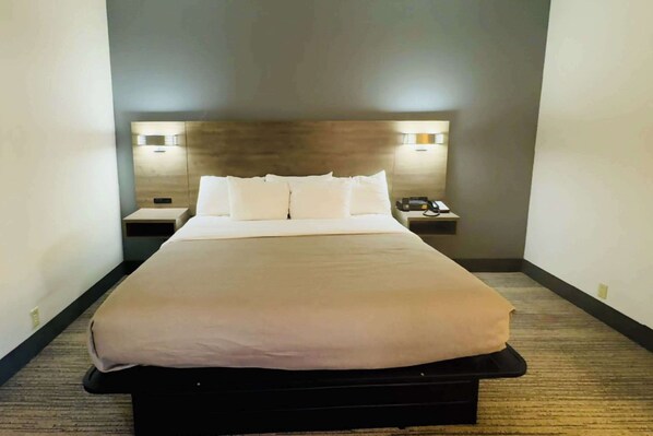 A neatly made bed with a beige bedspread and white pillows, a bedside table with a lamp, and a wall-mounted light fixture.