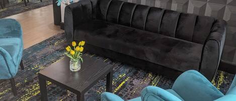 A modern living area with a black sofa, two blue armchairs, a small table with a vase of yellow flowers, and a patterned carpet.