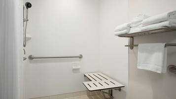 A shower with a handheld showerhead, a grab bar, and a small step for accessibility. There are white towels stacked on a shelf.