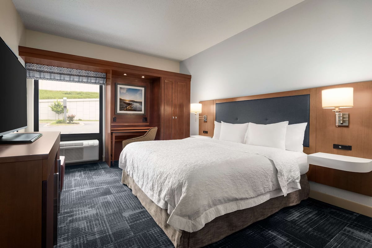 A hotel room with a large bed, a desk, a chair, a window with a view, and a wall-mounted lamp.